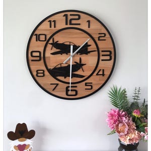 May include: Round wooden wall clock with black numerals and hands. The clock face features two black airplane silhouettes. The clock is surrounded by a black frame. The clock is on a light brown surface.