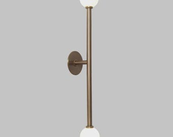 Puede incluir: Una lámpara de pared moderna con un diseño vertical. Cuenta con un marco de metal de color bronce con dos bombillas esféricas de vidrio blanco, una en la parte superior y otra en la inferior. El accesorio está montado sobre una base circular.