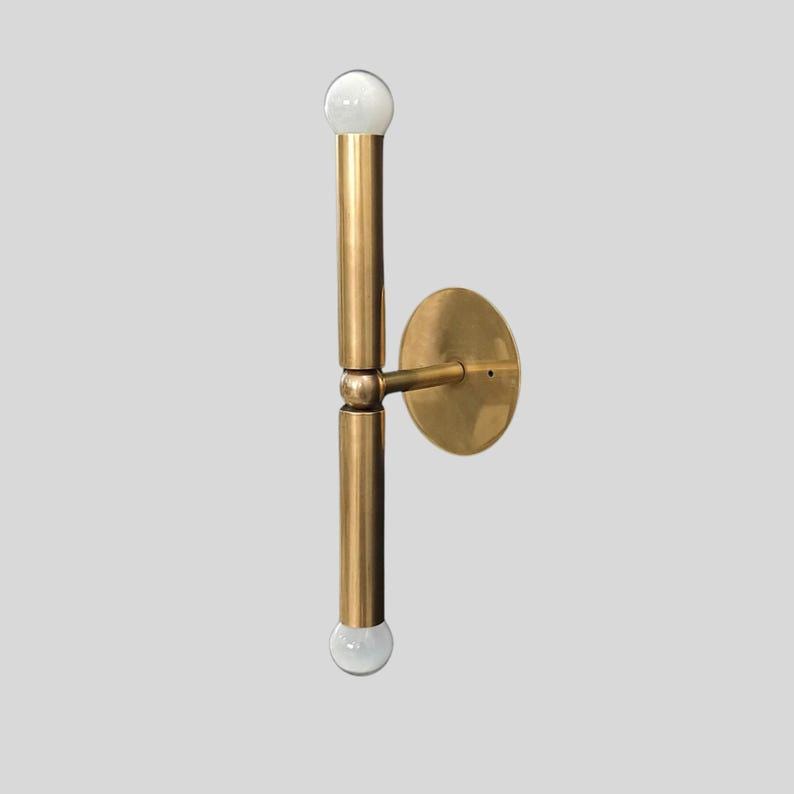 Puede incluir: Una l&aacute;mpara de pared de lat&oacute;n con un dise&ntilde;o vertical. La l&aacute;mpara tiene dos brazos cil&iacute;ndricos con una bombilla esf&eacute;rica en cada extremo. Una placa circular est&aacute; fijada a la pared y los brazos est&aacute;n conectados a la placa.