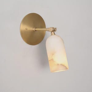 Può includere: Applique da parete con base rotonda color ottone e braccio orientabile. La lampada ha un paralume cilindrico in materiale traslucido bianco sporco con venature marroni.