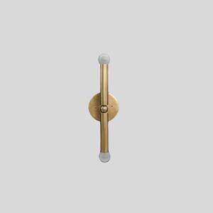 Puede incluir: Aplique de pared de lat&oacute;n con dos accesorios de iluminaci&oacute;n verticales y una base circular. Cada accesorio tiene una peque&ntilde;a bombilla redonda y esmerilada en el extremo. El aplique est&aacute; montado sobre un fondo gris claro.