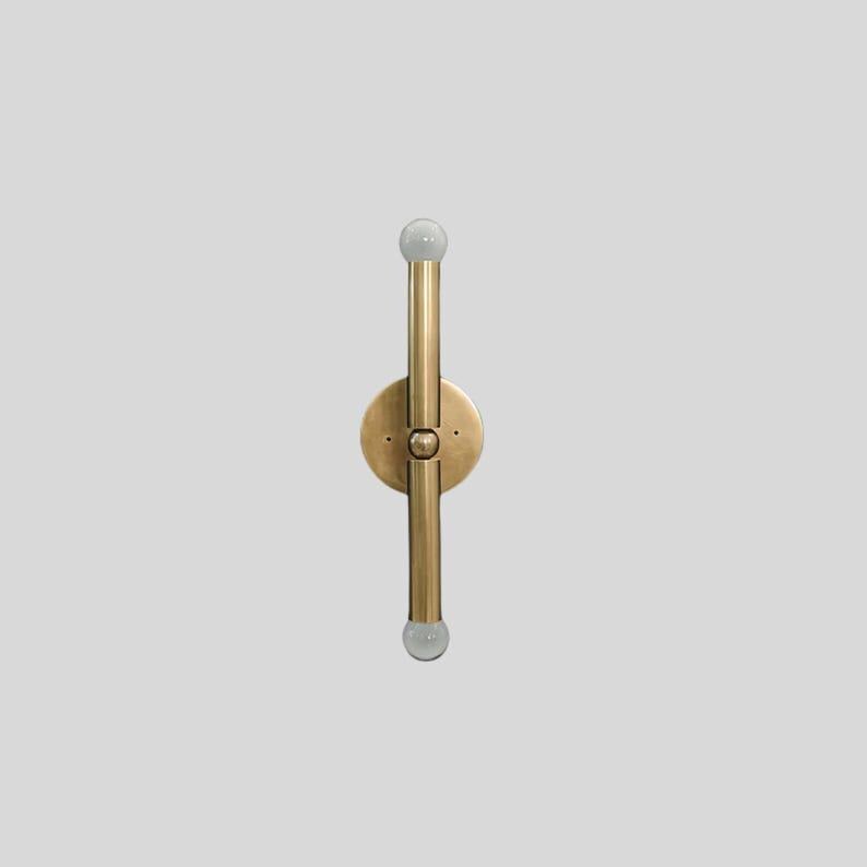 Puede incluir: Aplique de pared de lat&oacute;n con dos accesorios de iluminaci&oacute;n verticales y una base circular. Cada accesorio tiene una peque&ntilde;a bombilla redonda y esmerilada en el extremo. El aplique est&aacute; montado sobre un fondo gris claro.