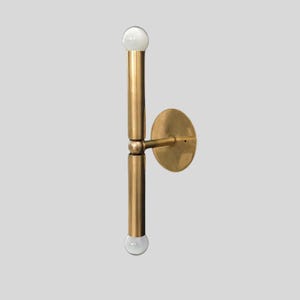 Puede incluir: Una l&aacute;mpara de pared de lat&oacute;n con un dise&ntilde;o vertical. La l&aacute;mpara tiene dos brazos cil&iacute;ndricos con una bombilla esf&eacute;rica en cada extremo. Una placa circular est&aacute; fijada a la pared y los brazos est&aacute;n conectados a la placa.