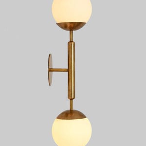 May include: A contemporary wall light with a brass finish. It has two spherical, frosted glass globes, one above and one below, joined by a vertical rod. A circular plate is fixed to the wall. The light fixture is designed for indoor use.