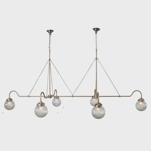 Könnte beinhalten: Eine mehrflammige Pendelleuchte in Bronzetönen mit sechs kugelförmigen, gerippten Glasschirmen. Die Leuchte hat ein lineares Design mit gebogenen Armen und einer Kettenaufhängung. Der Gesamtstil ist vintage und elegant, passend für ein Esszimmer oder eine Küche.