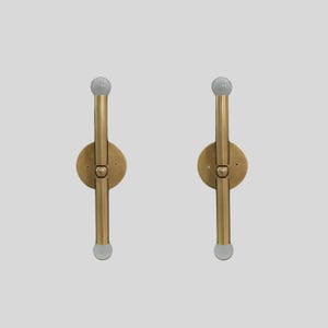 Puede incluir: Dos apliques de pared de lat&oacute;n con un dise&ntilde;o minimalista. Cada aplique presenta una base circular, una varilla vertical y un globo de vidrio esmerilado en la parte superior e inferior. Los apliques est&aacute;n montados sobre un fondo gris claro.