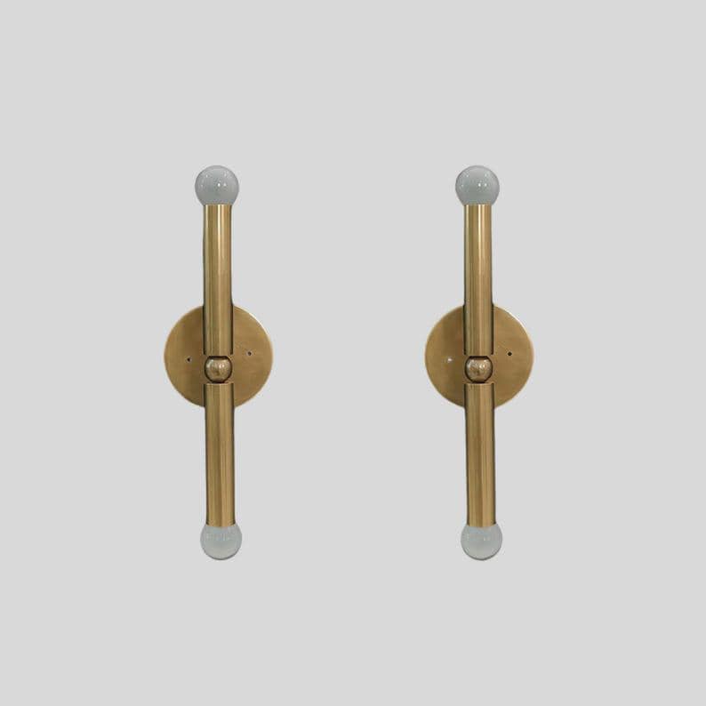 Puede incluir: Dos apliques de pared de lat&oacute;n con un dise&ntilde;o minimalista. Cada aplique presenta una base circular, una varilla vertical y un globo de vidrio esmerilado en la parte superior e inferior. Los apliques est&aacute;n montados sobre un fondo gris claro.