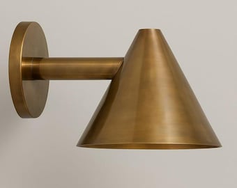 May include: A brass wall sconce with a conical shade and a circular backplate. The sconce has a straight arm connecting the shade to the backplate. The finish is a warm, metallic gold.