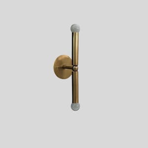 Puede incluir: Aplique de pared moderno en tono lat&oacute;n con base circular y dos luminarias verticales. Cada luminaria tiene un dise&ntilde;o cil&iacute;ndrico y est&aacute; rematada con una peque&ntilde;a bombilla esf&eacute;rica blanca. Dise&ntilde;ado para iluminaci&oacute;n interior.