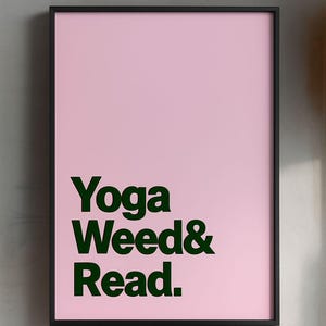May include: A framed pink poster with the words "Yoga Weed & Read" in dark green lettering. The poster has a black frame and is displayed against a light gray wall.