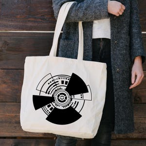 May include: Cream-colored tote bag featuring a bold black geometric design. The design is a circular pattern with segmented shapes, lines, and dots. The bag has long handles for carrying over the shoulder.