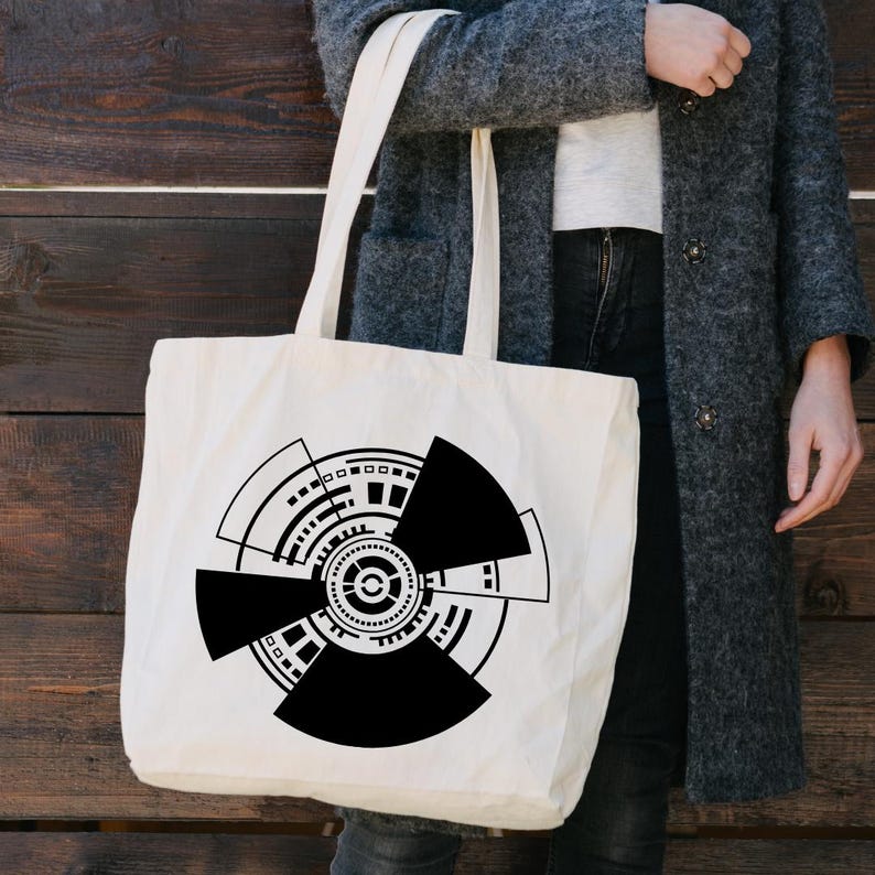 May include: Cream-colored tote bag featuring a bold black geometric design. The design is a circular pattern with segmented shapes, lines, and dots. The bag has long handles for carrying over the shoulder.