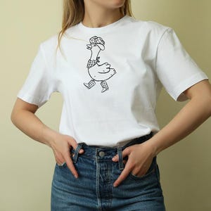 以下が含まれることがあります： 帽子、スカーフ、靴を身に着けたガチョウの白黒イラストが特徴の白いTシャツ。ガチョウは歩いている様子が描かれています。このシャツは柔らかい素材でできており、カジュアルウェアに適しています。