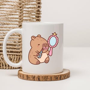 Op de afbeelding: Witte keramische mok met een cartoon capibara die lippenstift aanbrengt en in een roze handspiegel kijkt. De capibara is lichtbruin met een lichtbeige buik. De mok staat op een houten onderzetter.