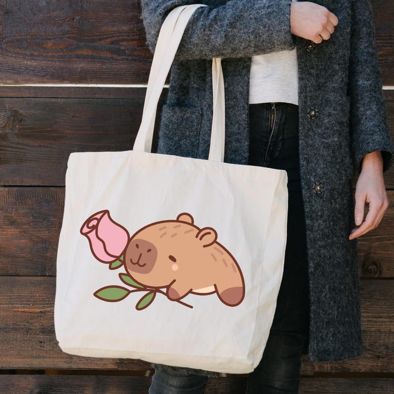 Op de afbeelding: Cr&egrave;mekleurige draagtas met een cartoon capibara en een roze roos. De capibara is lichtbruin met een lachend gezicht. De roos is roze met groene bladeren. De tas heeft lange handvatten en is gemaakt van een stevige stof.