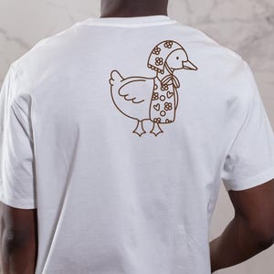 以下が含まれることがあります： 白いTシャツ。茶色の線画で、花柄とハート柄の帽子とスカーフを身につけたアヒルのイラスト。アヒルは右向き。Tシャツは無地で、半袖です。