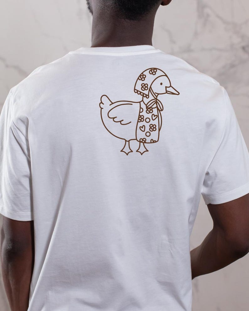 以下が含まれることがあります： 白いTシャツ。茶色の線画で、花柄とハート柄の帽子とスカーフを身につけたアヒルのイラスト。アヒルは右向き。Tシャツは無地で、半袖です。
