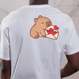 Op de afbeelding: Wit t-shirt met een cartoon illustratie van een bruine beer die een envelop met rode harten vasthoudt. Het ontwerp is gecentreerd op de achterkant van het shirt en heeft een schattig en romantisch thema.