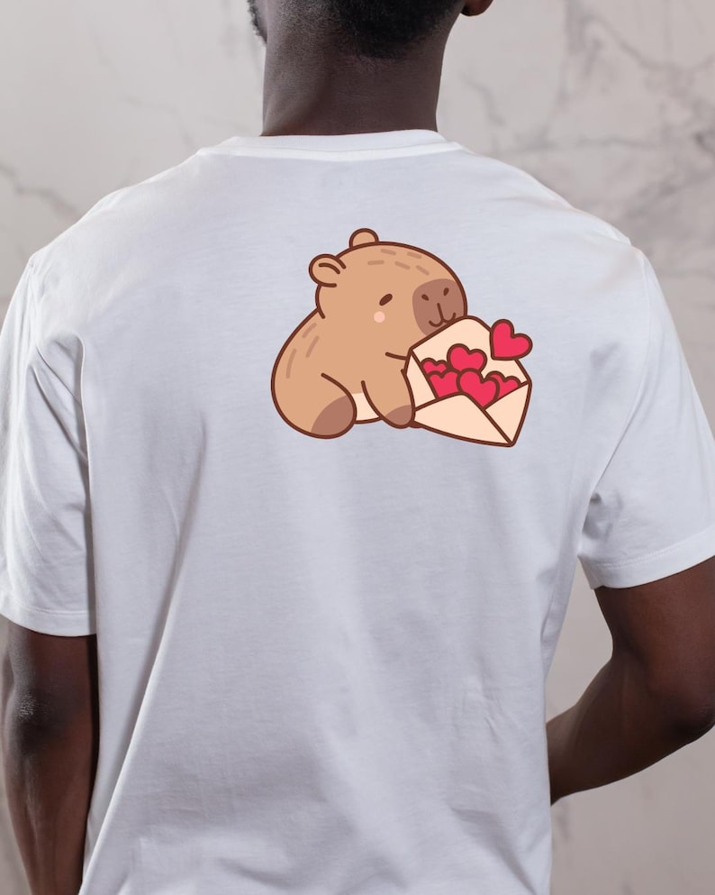 Op de afbeelding: Wit t-shirt met een cartoon illustratie van een bruine beer die een envelop met rode harten vasthoudt. Het ontwerp is gecentreerd op de achterkant van het shirt en heeft een schattig en romantisch thema.