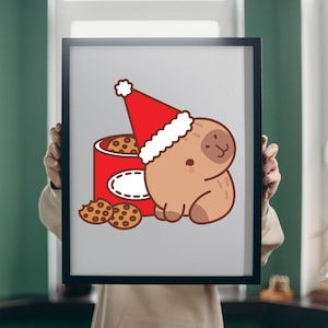 Op de afbeelding: Een ingelijste print met een cartoon capibara met een kerstmuts, met een rode koektrommel en chocolate chip cookies. De capibara is lichtbruin, de hoed is rood met witte rand, en de koekjes zijn bruin.