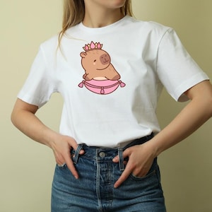 Op de afbeelding: Wit T-shirt met een cartoon capibara met een roze kroon en rustend op een roze kussen. De capibara is lichtbruin met een klein, lachend gezicht. Het kussen heeft een decoratieve rand. Het shirt is een klassieke ronde hals.