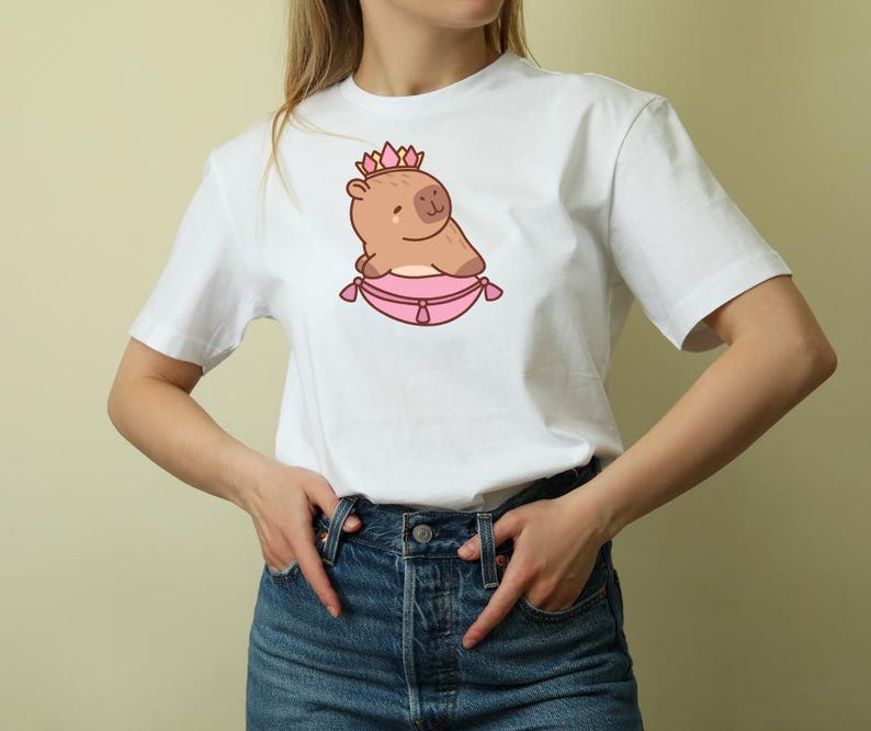 Op de afbeelding: Wit T-shirt met een cartoon capibara met een roze kroon en rustend op een roze kussen. De capibara is lichtbruin met een klein, lachend gezicht. Het kussen heeft een decoratieve rand. Het shirt is een klassieke ronde hals.