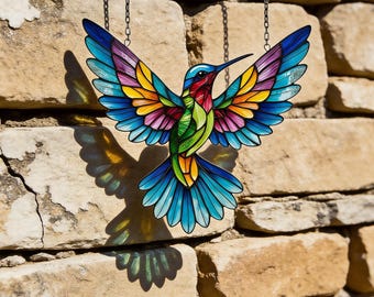 Op de afbeelding: Een kleurrijke kolibrie-zonnevanger met uitgespreide vleugels, hangend aan een ketting. De vogel heeft levendige kleuren blauw, groen, geel, roze en paars. De zonnevanger wordt getoond tegen een getextureerde stenen muur, die een gedetailleerde schaduw werpt.