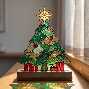 Könnte beinhalten: Eine Weihnachtsbaumlampe aus Buntglas mit strukturierten grünen und goldenen Glaselementen, roten runden Verzierungen und roten Geschenken am Fuß. Der Baum wird von einem leuchtenden Stern gekrönt und steht auf einem rechteckigen Holzsockel.