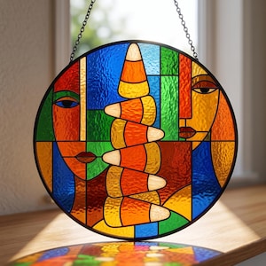 May include: A circular stained glass suncatcher featuring two stylized faces in profile facing a central vertical stack of candy corn. The piece uses vibrant red, blue, green, yellow, and orange glass segments held together by a black metal frame.