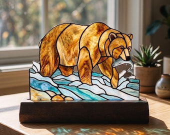 Op de afbeelding: Een glas-in-loodpaneel met een bruine beer die een vis vangt in een blauw-witte rivier. De beer is gemaakt van amberkleurige glasstukken, terwijl het water wervelend blauw en wit glas toont op een donkerhouten voetstuk.