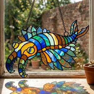 May include: A vibrant stained glass artwork of a sea creature, suspended by a chain. The design incorporates blue, green, orange, and gold hues. The piece is displayed in front of a window, showcasing its intricate details.