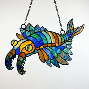May include: A stained glass artwork of a colourful, stylised sea creature. The piece features blues, greens, oranges, and yellows, with detailed patterns. It hangs from a black chain, ideal for window or wall decoration. The artwork is approximately 30 cm in length.