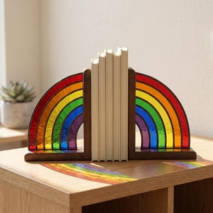 Puede incluir: Un par de sujetalibros en forma de L hechos de madera marrón oscura, cada uno con una pieza de vidrio tintado semicircular con un vibrante patrón de arcoíris en rojo, naranja, amarillo, verde, azul y morado, sosteniendo cuatro libros pequeños.