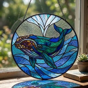 Könnte beinhalten: Rundes Buntglasfenster mit einer Wal-Darstellung in blau- und türkisfarbenem Wasser. Der Wal hat einen detaillierten Kopf mit Braun-, Lila- und Goldtönen. Das Kunstwerk ist schwarz gerahmt und wird von einer Kette gehalten.