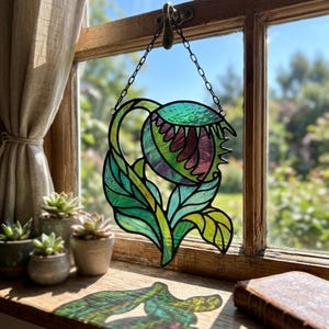May include: Stained glass art piece depicting a stylized plant with a large, open flower head and leaves. The piece features shades of green, purple, and yellow, with a black outline. It hangs from a chain in front of a window.