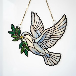 Puede incluir: Vidriera de una paloma en vuelo, sosteniendo una rama de olivo en su pico. La paloma est&aacute; hecha de vidrio blanco, azul y transparente con contornos negros. Cuelga de una cadena de lat&oacute;n, un s&iacute;mbolo de paz.