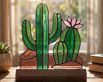 Op de afbeelding: Een cactussculptuur van glas-in-lood met levendige groene glaspanelen en donkere metalen soldeerverbindingen. Het object bevat een hoge centrale cactus, een kleinere cactus aan de zijkant en een lichtroze bloem op een houten voet.