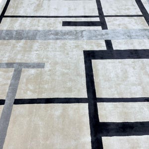 May include: A large, rectangular area rug with a geometric design. The rug features a neutral beige background with a pattern of black and gray lines forming rectangular shapes. The rug has a soft, plush texture.