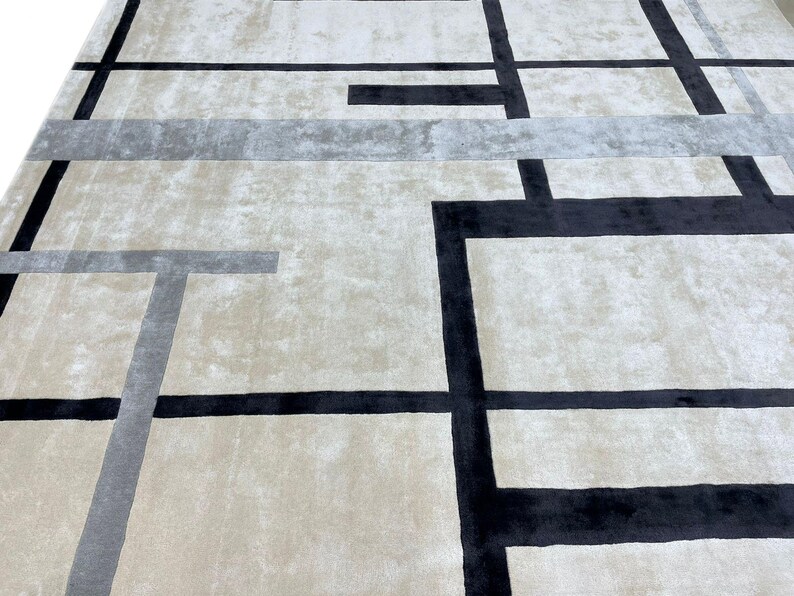 May include: A large, rectangular area rug with a geometric design. The rug features a neutral beige background with a pattern of black and gray lines forming rectangular shapes. The rug has a soft, plush texture.