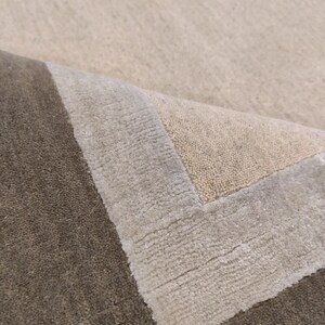 May include: A close-up of a beige and brown area rug. The rug has a textured surface with a border in shades of beige and gray. The corner of the rug is folded over, revealing the underside.