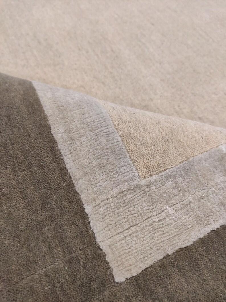 May include: A close-up of a beige and brown area rug. The rug has a textured surface with a border in shades of beige and gray. The corner of the rug is folded over, revealing the underside.