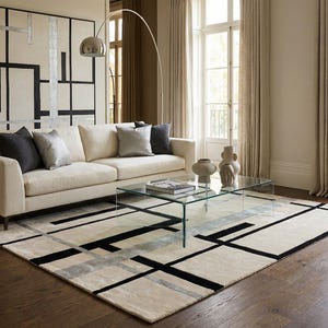 May include: A modern living room featuring a cream-colored sofa with decorative pillows, a glass coffee table, and a geometric rug with black and gray lines. A large abstract painting hangs on the wall, and a silver arc lamp provides lighting.
