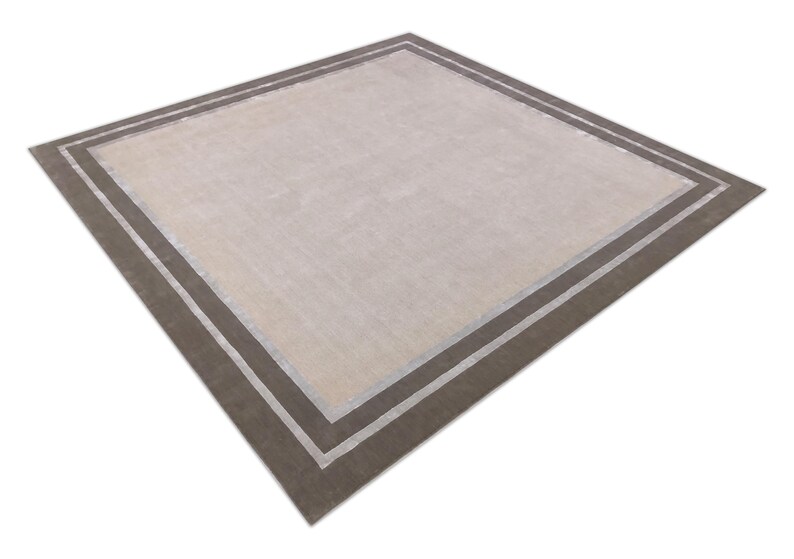 May include: A square area rug with a neutral beige center and a dark brown border. The border is accented with a thin, light-colored stripe. The rug has a simple, modern design.