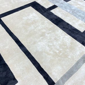 May include: A close-up view of a modern area rug with a geometric design. The rug features a cream-colored background with bold black and gray rectangular and linear patterns. The rug's texture is soft and plush.