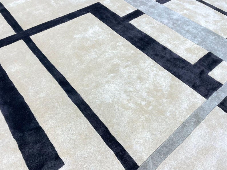 May include: A close-up view of a modern area rug with a geometric design. The rug features a cream-colored background with bold black and gray rectangular and linear patterns. The rug's texture is soft and plush.