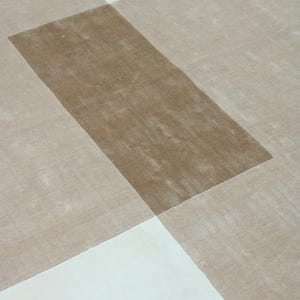 Pu&ograve; includere: Un tappeto moderno con un design geometrico. Il tappeto presenta una sezione rettangolare centrale di colore marrone medio, sovrapposta a un motivo di sezioni beige e bianche. Il design crea un effetto astratto a strati.