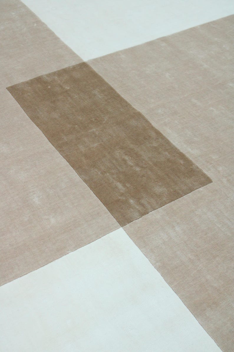 Pu&ograve; includere: Un tappeto moderno con un design geometrico. Il tappeto presenta una sezione rettangolare centrale di colore marrone medio, sovrapposta a un motivo di sezioni beige e bianche. Il design crea un effetto astratto a strati.