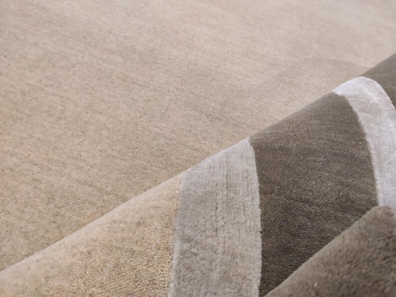 May include: Close-up of a rolled-up area rug with a neutral color palette. The rug features a beige background with a design of vertical stripes in shades of gray and brown. The texture of the rug is visible.