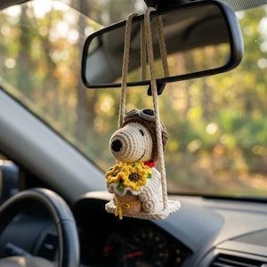 May include: Crocheted car charm featuring a Snoopy-like dog wearing a pilot's cap and goggles, holding a basket of sunflowers. The charm hangs from the rearview mirror inside a car, with a blurred forest background.