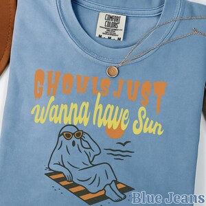May include: A light blue t-shirt featuring a graphic of a ghost wearing sunglasses while lounging on a striped beach towel. The text reads GHOULS JUST wanna have Sun. The shirt has a Comfort Colors label at the neck and a small gold pendant necklace.